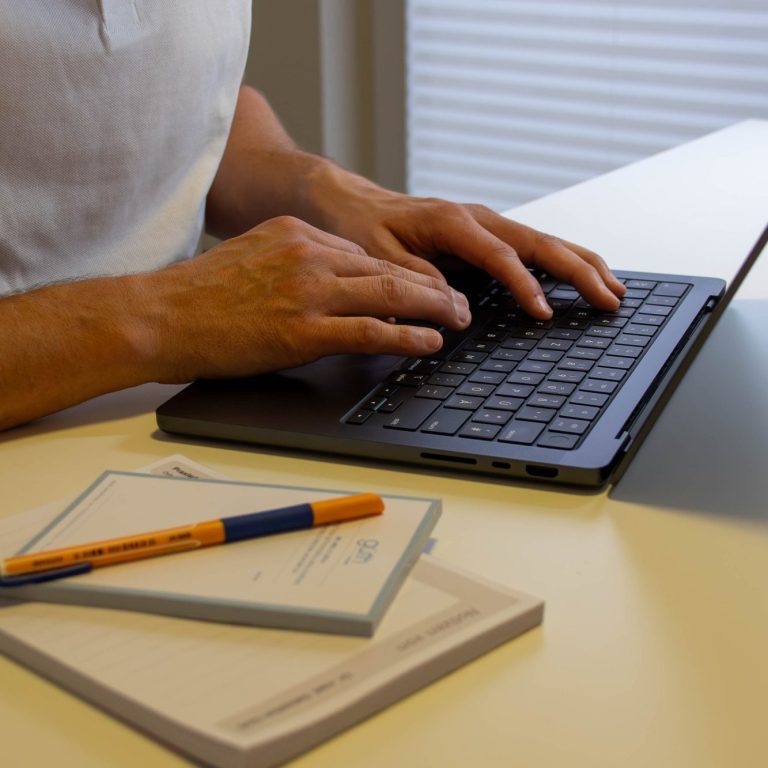 Dr. Güth tippt auf dem Laptop, Rezeptblock und ein Stift liegen auf dem Tisch.